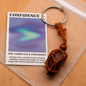 May include: A red tiger's eye stone wrapped in brown cord and attached to a silver key ring. The stone is in a white organza bag. The card reads "CONFIDENCE" and "RED TIGERS EYE & CONFIDENCE". Red Tigers Eye a powerful stone for boosting confidence and personal strength. It's vibrant, energy helps enhance self esteem and courage.