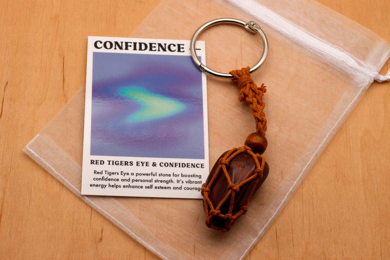 May include: A red tiger's eye stone wrapped in brown cord and attached to a silver key ring. The stone is in a white organza bag. The card reads "CONFIDENCE" and "RED TIGERS EYE & CONFIDENCE". Red Tigers Eye a powerful stone for boosting confidence and personal strength. It's vibrant, energy helps enhance self esteem and courage.