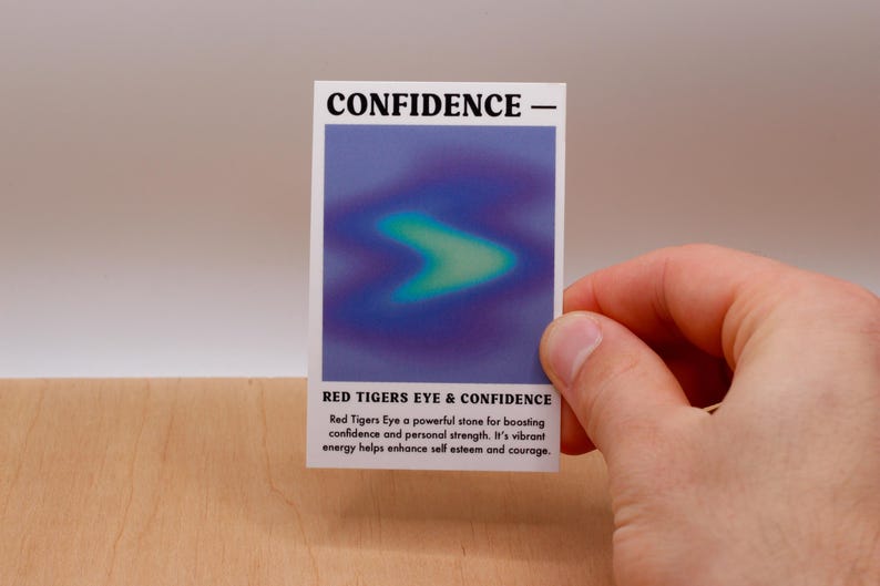 May include: A white card with the text "CONFIDENCE -" at the top and "RED TIGERS EYE & CONFIDENCE" at the bottom. The card has a blue and green gradient image in the center. The text below the image reads: "Red Tigers Eye a powerful stone for boasting confidence and personal strength. It's vibrant energy helps enhance self esteem and courage."