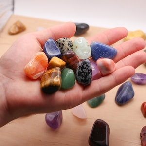 May include: A collection of colourful tumbled stones, including amethyst, tiger's eye, and jasper, in a hand. The stones are arranged in a pile on a wooden surface.