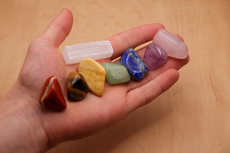 May include: A hand holding a set of seven chakra healing crystals, including selenite, tiger's eye, citrine, aventurine, lapis lazuli, amethyst, and rose quartz.