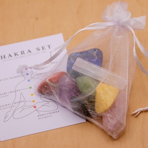 May include: A set of seven chakra crystals in a white mesh bag. The crystals are arranged in a rainbow order, with the root chakra crystal at the bottom and the crown chakra crystal at the top. The bag is tied with a white ribbon. A card with a diagram of the chakras is also included.