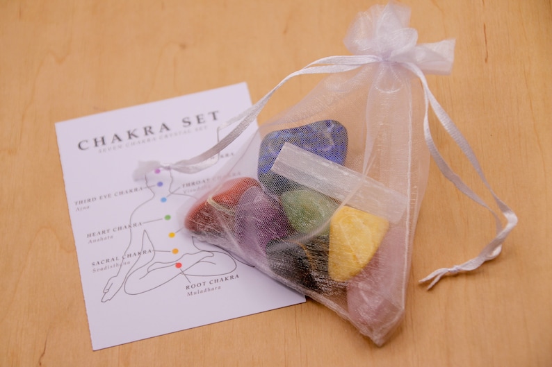May include: A set of seven chakra crystals in a white mesh bag. The crystals are arranged in a rainbow order, with the root chakra crystal at the bottom and the crown chakra crystal at the top. The bag is tied with a white ribbon. A card with a diagram of the chakras is also included.
