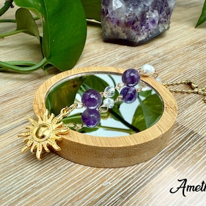 Puede incluir: Un dije dorado con forma de sol y un collar con dos cuentas de amatista moradas se exhiben sobre un peque&ntilde;o espejo redondo. El espejo est&aacute; enmarcado en madera. La palabra "Amethyst" est&aacute; escrita en cursiva.