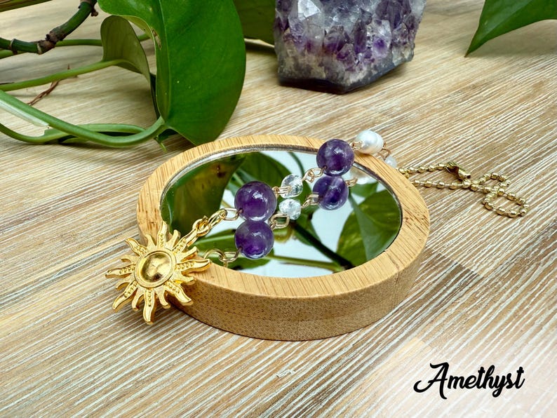 Puede incluir: Un dije dorado con forma de sol y un collar con dos cuentas de amatista moradas se exhiben sobre un peque&ntilde;o espejo redondo. El espejo est&aacute; enmarcado en madera. La palabra "Amethyst" est&aacute; escrita en cursiva.