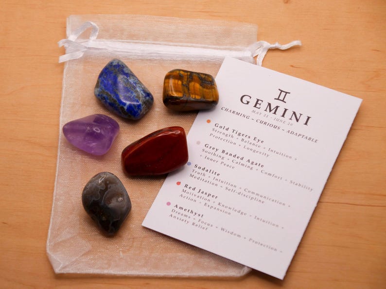 Peut inclure: Un ensemble de sept cristaux dans un sac en organza blanc avec une carte qui indique "GEMINI II MAY 21 - JUNE 21 CHARMING - CURIOUS - ADAPTABLE" et liste les cristaux et leurs propri&eacute;t&eacute;s. Les cristaux sont l'am&eacute;thyste, le jaspe rouge, la sodalite, l'agate ray&eacute;e grise, l'&oelig;il de tigre dor&eacute; et deux cristaux bleus.