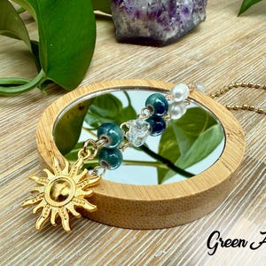 Puede incluir: Un dije de sol dorado cuelga de una cadena, con cuentas verdes y azules y un peque&ntilde;o espejo. El texto "Green Agate" est&aacute; escrito en escritura negra. El espejo est&aacute; enmarcado en un marco de madera redondo.
