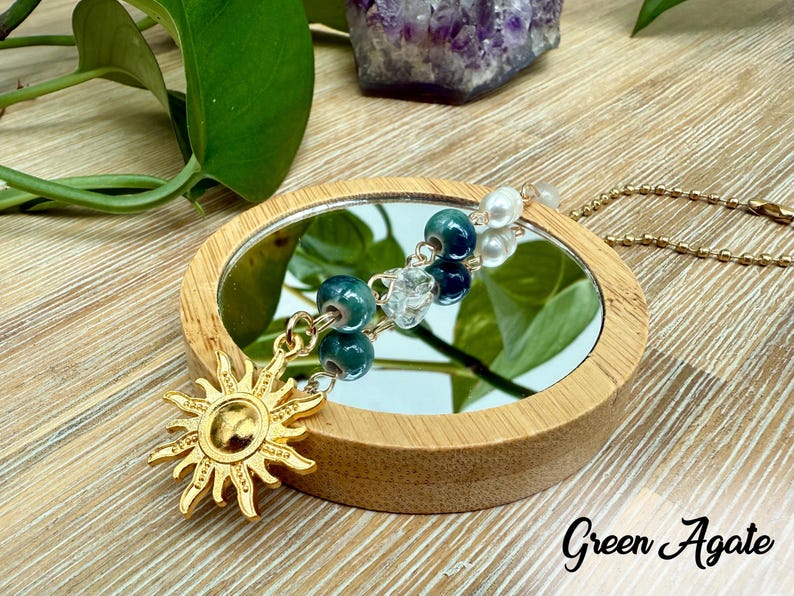 Puede incluir: Un dije de sol dorado cuelga de una cadena, con cuentas verdes y azules y un peque&ntilde;o espejo. El texto "Green Agate" est&aacute; escrito en escritura negra. El espejo est&aacute; enmarcado en un marco de madera redondo.