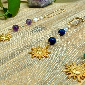 Puede incluir: Colgantes dorados con forma de sol y cadenas de cuentas. Los colgantes presentan un dise&ntilde;o de sol. Las cadenas est&aacute;n adornadas con varias cuentas, incluyendo amatista, lapisl&aacute;zuli azul y perlas blancas. Los colgantes se exhiben sobre una superficie de madera.