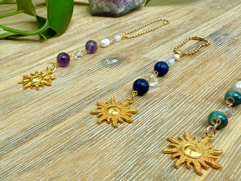 Puede incluir: Colgantes dorados con forma de sol y cadenas de cuentas. Los colgantes presentan un dise&ntilde;o de sol. Las cadenas est&aacute;n adornadas con varias cuentas, incluyendo amatista, lapisl&aacute;zuli azul y perlas blancas. Los colgantes se exhiben sobre una superficie de madera.