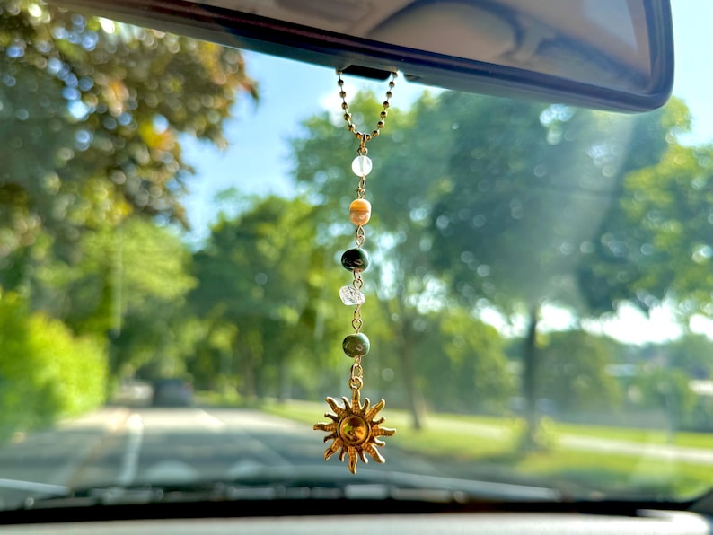 Puede incluir: Un adorno de coche dorado con un colgante de sol y una cadena de cuentas de colores. Las cuentas incluyen piedras transparentes, verdes y beige. El adorno cuelga del espejo retrovisor de un coche, con un fondo borroso de &aacute;rboles y una carretera.