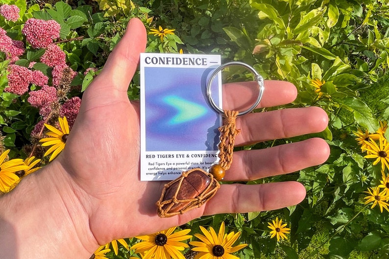 May include: A red tiger's eye stone wrapped in brown leather cord with a wooden bead and a silver key ring. The stone is attached to a card that reads "CONFIDENCE - Red Tiger's Eye & Confidence".