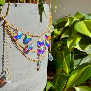 Puede incluir: Una cadena dorada con múltiples hebras y colgantes de cristal iridiscente. La cadena está unida a un gancho y cuelga frente a una maceta de hormigón con plantas verdes.
