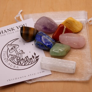 May include: A set of seven tumbled chakra stones in a white organza bag. The stones are amethyst, tiger's eye, lapis lazuli, red jasper, rose quartz, aventurine, and selenite. The bag has a drawstring closure and is tied with a white ribbon. The stones are arranged in a rainbow pattern. The bag is sitting on a wooden surface. The image also includes a thank you card with a mushroom and crystal design.