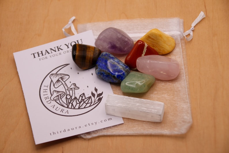 May include: A set of seven tumbled chakra stones in a white organza bag. The stones are amethyst, tiger's eye, lapis lazuli, red jasper, rose quartz, aventurine, and selenite. The bag has a drawstring closure and is tied with a white ribbon. The stones are arranged in a rainbow pattern. The bag is sitting on a wooden surface. The image also includes a thank you card with a mushroom and crystal design.