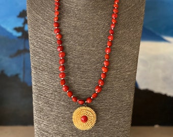 May include: A red seed bead necklace with a large, round, wooden pendant. The pendant is a flower shape with a red center. The necklace is displayed on a gray mannequin.