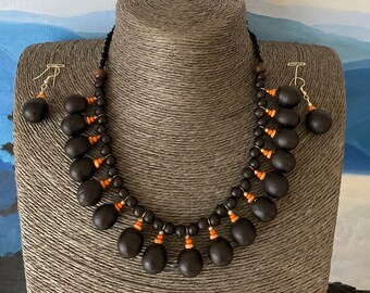 May include: A black seed necklace and matching earrings with orange beads. The necklace is made of a string of black beads with a row of black seeds hanging down from the bottom. The earrings are simple black seed drops.