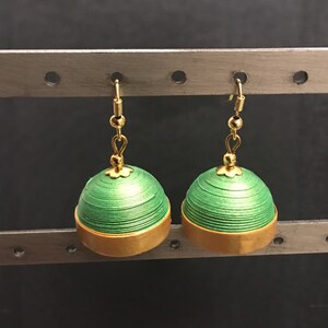 May include: A pair of green and gold dangle earrings. The earrings are made of paper and have a layered, textured design. The earrings are hanging from a metal stand.