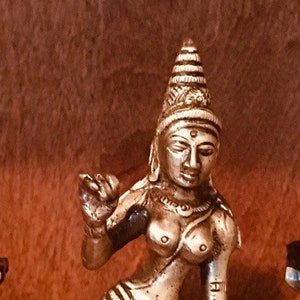 May include: A silver-colored metal statue of a female figure with a crown on her head. The statue is standing on a round base with intricate designs. The figure is wearing a long skirt and has her arms raised in a gesture of prayer.