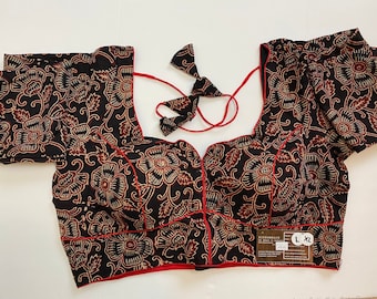 May include: Black blouse with a floral pattern in shades of brown and cream. The blouse has short sleeves, a scoop neckline, and red trim. It features a tie closure at the back and a tag with size information.