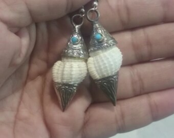 May include: A pair of silver earrings featuring white seashells and turquoise stones. The earrings are hanging from a person's hand.