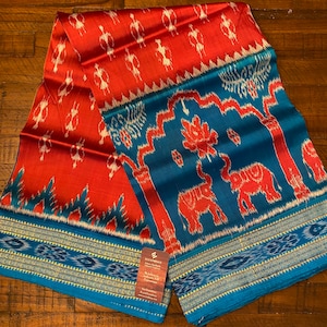 May include: A red and blue silk fabric with a traditional ikat pattern. The fabric features elephants and trees. The fabric has a blue and gold border. The fabric is labeled "Eco-Embrace".