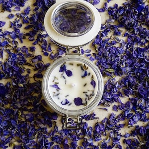May include: A white candle with dried lavender and purple flower petals in a clear glass jar with a white lid. The jar has a silver clasp.