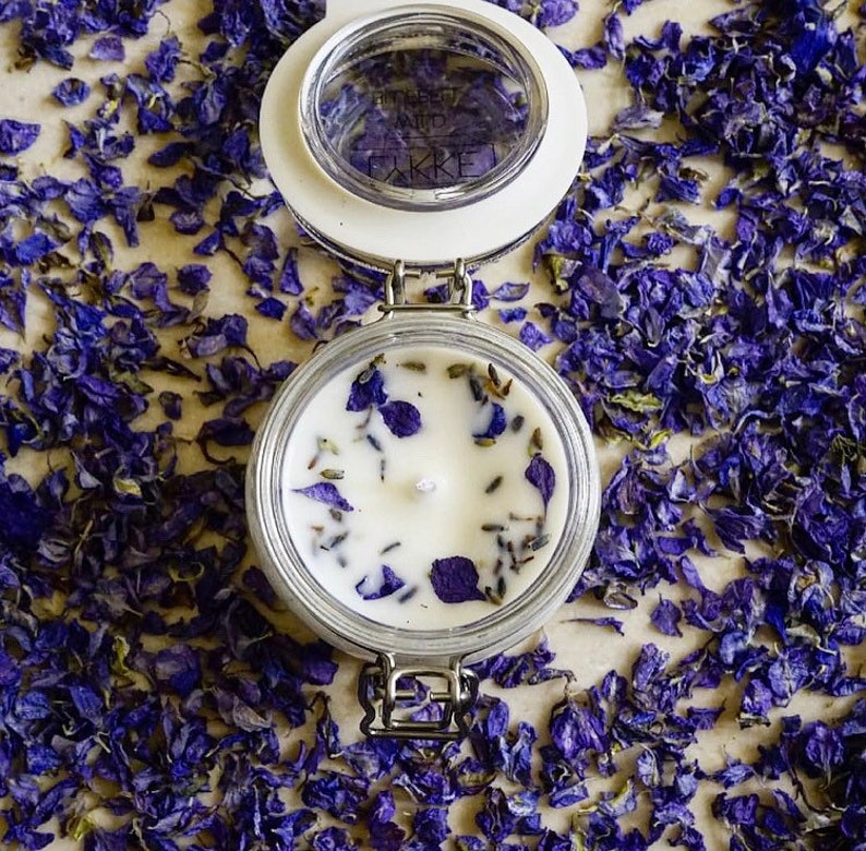 May include: A white candle with dried lavender and purple flower petals in a clear glass jar with a white lid. The jar has a silver clasp.