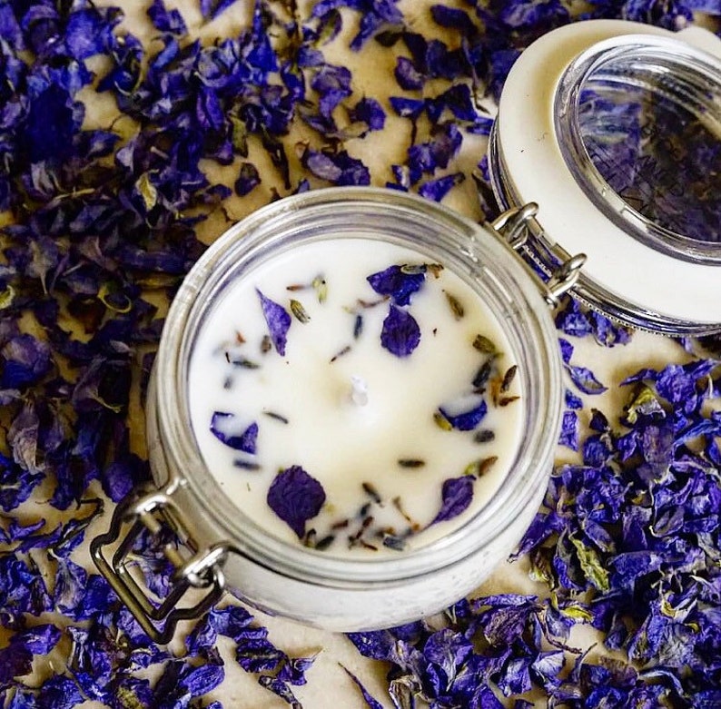 May include: A white soy candle with dried lavender and purple flower petals in a clear glass jar with a silver lid.