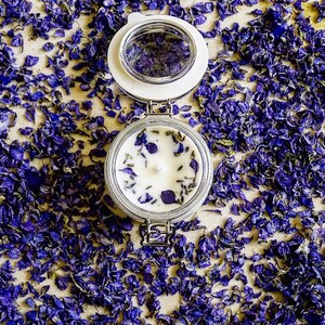 May include: A white candle with dried lavender and purple flower petals in a clear glass jar with a white lid. The jar is sitting on a surface covered in purple flower petals.
