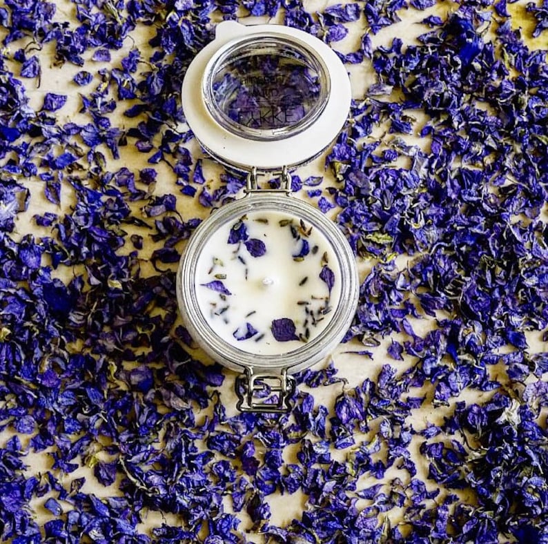 May include: A white candle with dried lavender and purple flower petals in a clear glass jar with a white lid. The jar is sitting on a surface covered in purple flower petals.