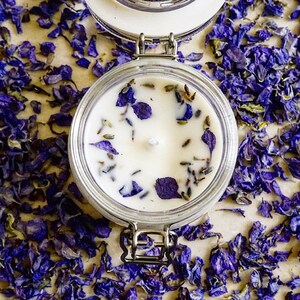 May include: A white candle with dried lavender and purple flower petals embedded in the wax. The candle is in a clear glass jar with a metal lid.