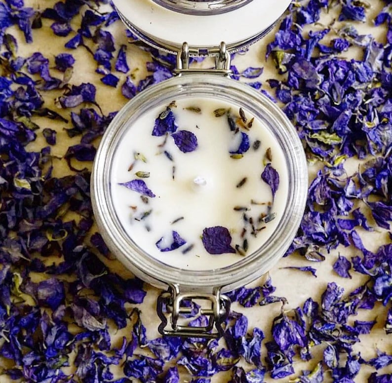 May include: A white candle with dried lavender and purple flower petals embedded in the wax. The candle is in a clear glass jar with a metal lid.