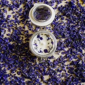 May include: A white candle with dried lavender and purple flower petals in a clear glass jar with a white lid. The jar is surrounded by purple flower petals.
