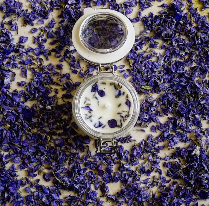 May include: A white candle with dried lavender and purple flower petals in a clear glass jar with a white lid. The jar is surrounded by purple flower petals.