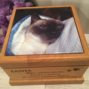 May include: A wooden box with a ceramic tile top featuring a photo of a Siamese cat. The box has an engraved inscription that reads "SASHA When someone you love becomes a memory the memory becomes a treasure. AUG. 19, 2018".