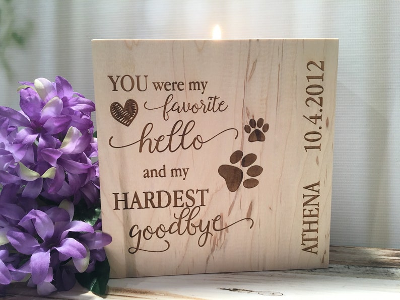 May include: A wooden memorial candle with the text "YOU were my favorite hello and my HARDEST goodbye" and two paw prints. The candle is engraved with the name "ATHENA" and the date "10.4.2012".