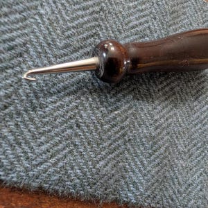 May include: A rug hooking tool with a dark brown wooden handle and a silver metal hook, lying on a grey herringbone fabric. The handle is polished, and the hook is curved. The fabric has a distinctive chevron pattern.