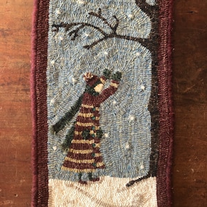 May include: A hand-hooked rug depicting a woman in a red and brown striped coat standing in the snow under a tree. The background is blue with white snowflakes.