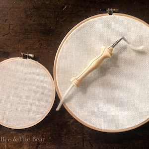 Op de afbeelding: Twee borduurringen met wit Aida-doek erover gespannen. De grotere ring heeft een houten borduurnaaldgereedschap erop liggen. De kleinere ring bevindt zich links van de grotere.