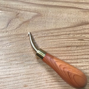 May include: A wood-handled leatherworking tool with a curved, brass-colored blade. The handle is a light brown color with visible wood grain. The tool is designed for shaping and cutting leather.