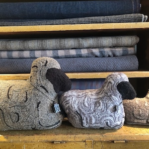 May include: Three gray and black sheep rug ornaments on a wooden shelf. The sheep are made of a textured fabric and have black faces and legs. The sheep are sitting on a wooden shelf with a yellow paint finish.