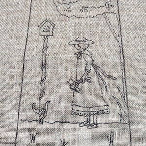 May include: Black and white cross-stitch embroidery on a beige fabric. The design features a woman in a dress and hat walking past a birdhouse on a pole.