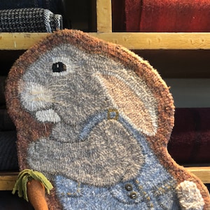 Puede incluir: Una alfombra marrón y gris con forma de conejo que lleva un mono azul. El conejo tiene la nariz blanca y bigotes. La alfombra está hecha de lana y tiene una superficie texturizada.