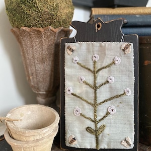 May include: A black wooden clipboard with a light green fabric panel featuring an embroidered floral design. The image also includes a moss-covered sphere, a decorative vase, and two small, aged paper pots. The text "2025 The Bee & The Bear" is at the bottom.