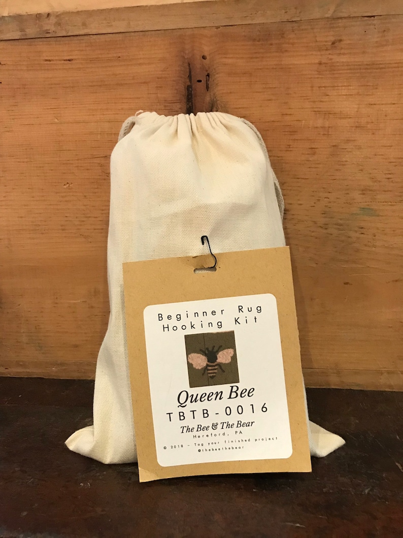 K&ouml;nnte beinhalten: Ein brauner Papierbeutel mit einem wei&szlig;en Beutel mit Kordelzug im Inneren. Der Beutel hat ein Etikett mit der Aufschrift "Beginner Rug Hooking Kit" und "Queen Bee TBTB-0016". Das Etikett enth&auml;lt auch ein kleines Bild einer Biene mit rosa Fl&uuml;geln.