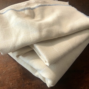 May include: Three pieces of beige Aida cloth folded together. The top piece has a light blue border.