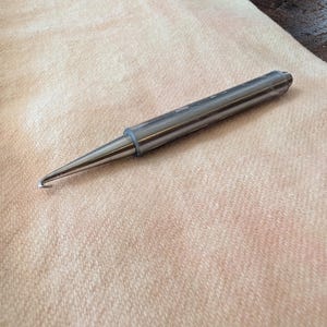 May include: A silver crochet hook with a curved end, lying on a light peach-colored fabric. The hook is cylindrical with a pointed tip and a small ring near the hook's curve. The hook is designed for yarn crafts.