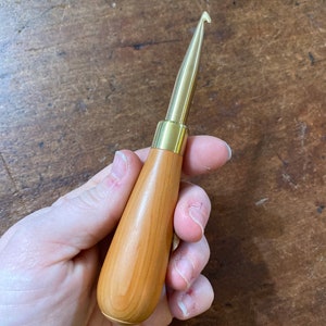 May include: A crochet hook featuring a smooth, light-toned wooden handle with a tapered shape and a polished gold-coloured metal hook tip. The tool has a cylindrical metal ferrule connecting the wooden handle to the metal hook component.