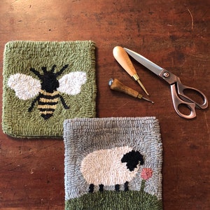 K&ouml;nnte beinhalten: Zwei handgestickte Woll-Teppich-H&auml;kel-Sets, eines mit einem Bienen-Design und das andere mit einem Schaf-Design. Die Sets befinden sich auf einer Holzoberfl&auml;che mit zwei Sticknadeln und einer Schere.